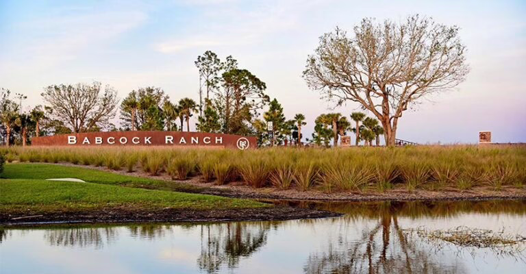 Entrance to Babcock Ranch featuring a large brick sign with white lettering, surrounded by tall grasses, palm trees, and a leafless tree, with a reflective pond in the foreground under a soft pastel sky.